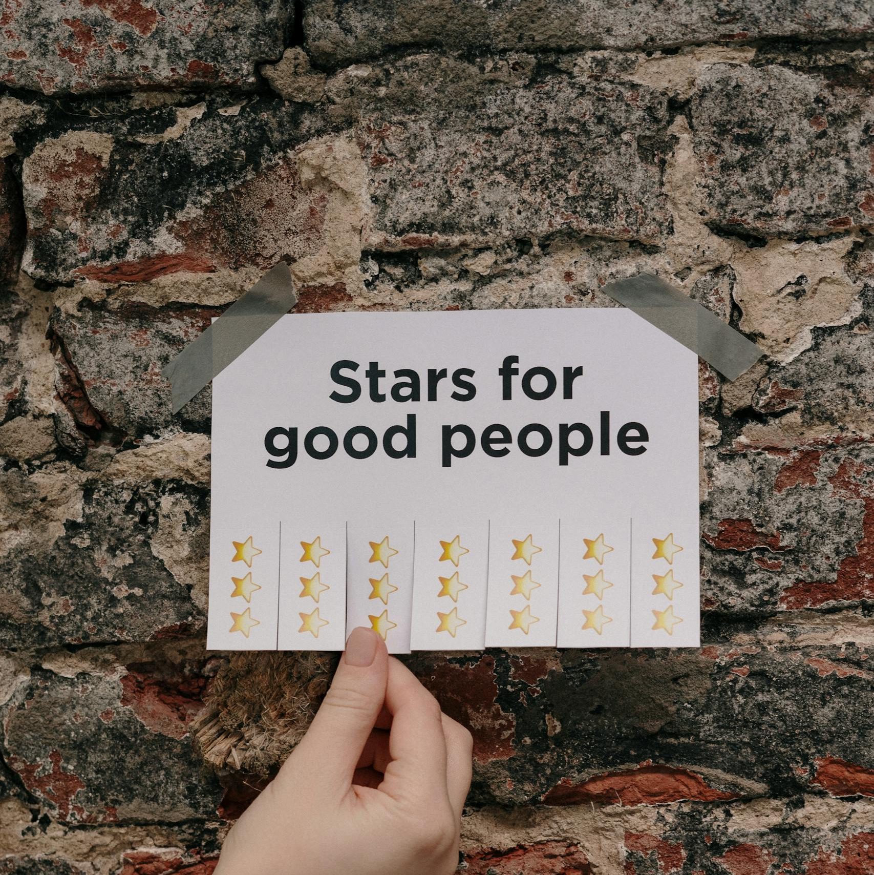 A close-up of a hand holding a printed paper with stars, taped to a rustic brick wall.